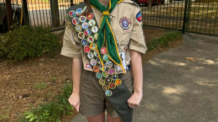 Allison in Scout uniform with a merit badge sash and green neckerchief, smiling on a garden path in front of a wrought-iron fence
