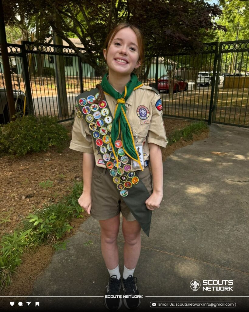 Allison in Scout uniform with a merit badge sash and green neckerchief, smiling on a garden path in front of a wrought-iron fence