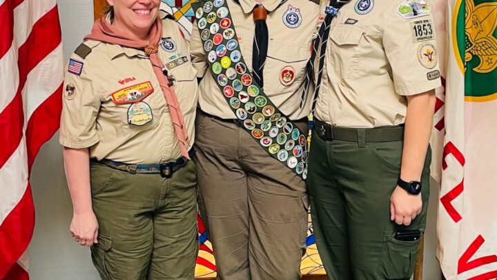 Eagle Scout Miaya in full merit badge sash standing with two Scout leaders in uniform in front of a stained-glass window