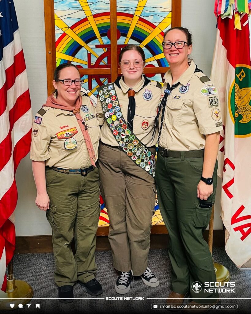 Eagle Scout Miaya in full merit badge sash standing with two Scout leaders in uniform in front of a stained-glass window