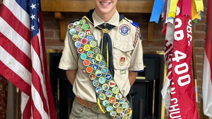 Lealond in Scout uniform and cap with a full merit badge sash, standing between the American flag and troop flags in front of a brick fireplace