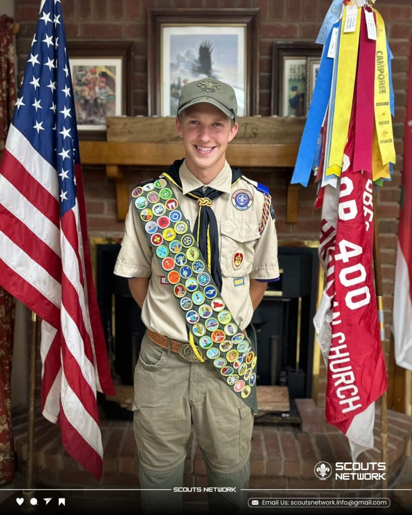 Lealond in Scout uniform and cap with a full merit badge sash, standing between the American flag and troop flags in front of a brick fireplace